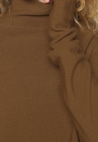 Brązowy, dzianinowy sweter z golfem i długim rękawem, o gładkiej fakturze i luźnym kroju, podkreślający ciepło i komfort.