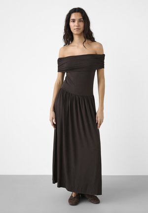 Donna in piedi che indossa un lungo vestito marrone scuro scollato e scarpe marroni su uno sfondo bianco uniforme.