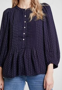 Blusa blu navy con motivi floreali bianchi, chiusura frontale con bottoni, dettagli plissettati e maniche a sbuffo; abbinata a jeans azzurri chiari.