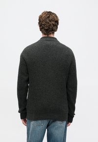 Ein Mann mit welligem, hellbraunem Haar trägt einen dunkelgrauen, strukturierten Pullover und blaue Jeans, während er mit dem Rücken zu einem schlichten, weißen Hintergrund steht.