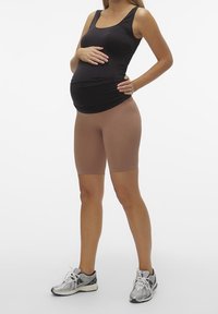 Černý bezrukávový top, nahé kompresní šortky a stříbrné sportovní boty. Modelka stojí s rukou na těhotenském břiše, neutrální pozadí.