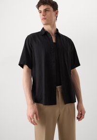 Maglietta nera a maniche corte in tessuto leggero con bottoni frontali e taschino sul petto, abbinata a pantaloni beige.