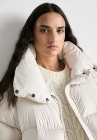 Napihnjena bela jakna z visokim ovratnikom, z gumbi in zadrgo, čez kremo pleteno jopico z gladko teksturo.