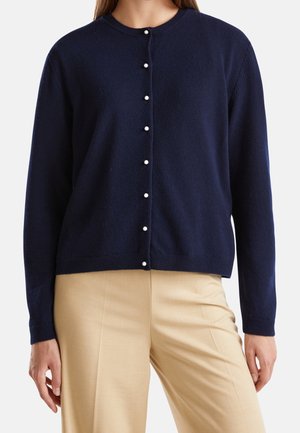 Marineblauwe cardigan met parelknopen gedragen over beige broek bij een persoon die voor een effen achtergrond staat.