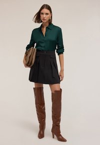 Cămașă de satin verde smarald, fustă mini plisată neagră și cizme cu toc înalt de culoare maronie până la coapse. Modelul ține o geantă de mână de culoare taupe.