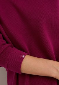 Haut à manches longues côtelé magenta avec un poignet retroussé. Présente une petite étiquette avec un logo en forme de cœur sur la manche. Texture lisse, coupe décontractée.