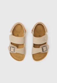 Μπεζ σανδάλια με δύο ρυθμιζόμενους ιμάντες που ασφαλίζουν με αγκράφες, κατασκευασμένα από μαλακό υλικό που μοιάζει με δέρμα, με καφέ σουέτ πάτο και ανάγλυφο λογότυπο.