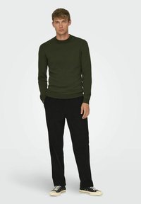 Dunkelgrüner strukturierter Pullover mit Rundhalsausschnitt, kombiniert mit schwarzen Hosen und schwarzen Turnschuhen, vor einem hellen Hintergrund stehend.