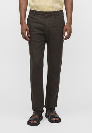 Homme portant un pantalon droit marron foncé, une chemise beige rentrée et des sandales noires, debout devant un fond uni.