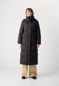 Γυναίκα που φοράει μακριό μαύρο καπιτονέ παλτό με ζώνη, μπεζ παντελόνια φαρδιάς γραμμής και μαύρα παπούτσια, που στέκεται μπροστά σε απλό λευκό φόντο.