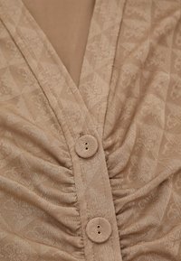 Beige fabric top featuring a textured diamond pattern, a V-neckline, gathered detailing, and two buttons at the front center.