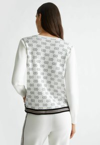 Maglione bianco a maglia con un motivo a logo grigio ripetuto. Presenta maniche lunghe, orlo a strisce nere e oro, e una schiena testurizzata.