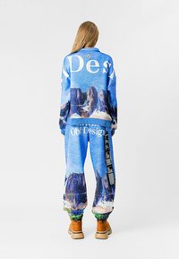 Chándal de color azul con un diseño de montañas, con una marca blanca en la chaqueta y los pantalones. El material parece texturizado; se lleva con botas marrón claro.