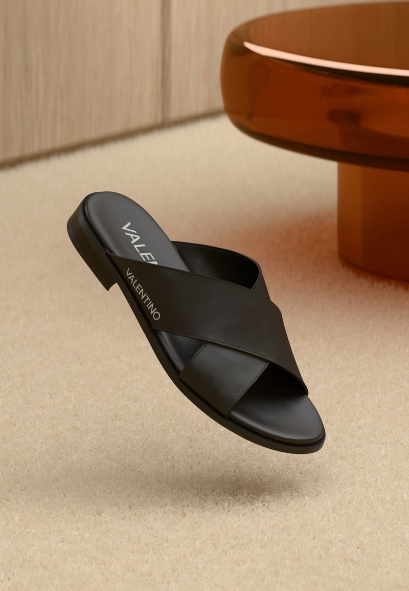 Svart lær slidesandal med kryssmønster og flat såle. Har merkevarelogo på innersålen. Plassert mot en myk beige bakgrunn.