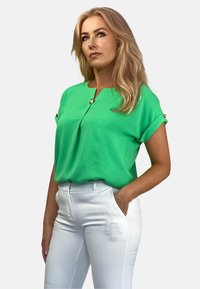 Zielona bluzka z krótkimi rękawami i dekoltem w kształcie litery V, z guzikiem jako detalem. Połączona z białymi spodniami, które mają gładką teksturę.