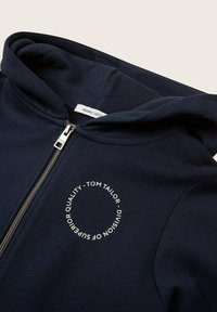 Dunkelblaues Kapuzenshirt aus Bio-Baumwolle, mit einem silbernen Reißverschluss und einem runden weißen Textlogo auf der Vorderseite.