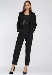 Czarny garnitur z krótką marynarką i spodniami z wysokim stanem, w połączeniu z czarnym topem zdobionym cekinami oraz czarnymi szpilkami. Gładka faktura tkaniny.
