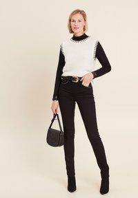 Stricktop in Weiß ohne Ärmel mit schwarzem Saum, übereinander getragen mit einem schwarzen Langarmshirt. Schwarze High-Waist-Jeans und eine kleine schwarze Handtasche.