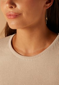 Gouden oorhangers met een kettingontwerp, met een staaf en bedel details. Het model draagt een lichtbeige geribbelde top met een ronde hals.