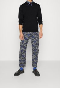 Pull noir en tricot, pantalon à motifs bleu-gris, chaussettes bleues et chaussures noires avec une sangle à accent rouge. Écharpe nouée autour du cou.