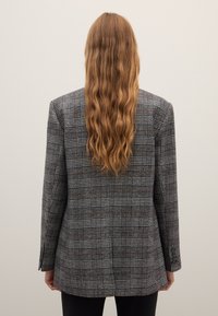 Grijze ruitenblazer met zwarte en bruine accenten, gestructureerd ontwerp, lange mouwen en manchetten met één knoop, gedragen over zwarte broek.