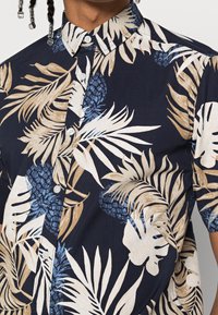 Camisetta a maniche corte con una base blu navy scura, caratterizzata da grandi motivi tropicali di foglie beige e blu e ananas; colletto con bottoni; tessuto morbido.