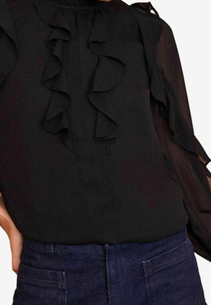 Blusa nera con dettagli a rouches sul davanti e sulle maniche, caratterizzata da un colletto alto e elementi in tessuto trasparente, abbinata a jeans in denim blu scuro.