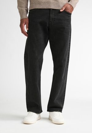Mann trägt schwarze Jeans, beigen Pullover und weiße Sneakers, steht mit einer Hand in der Tasche vor einfarbigem Hintergrund.