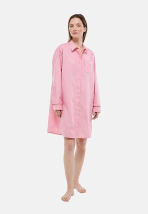 Femme debout pieds nus portant une robe chemise oversize à rayures verticales roses et blanches, avec des manches longues et une poche poitrine.