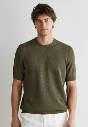 Man met kort bruin haar, gekleed in een olijfgroene, gestructureerde trui met korte mouwen en witte broek tegen een effen witte achtergrond.