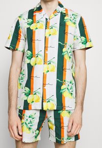 Conjunto de camisa de manga corta y pantalones cortos con estampado de limones, que presenta rayas verdes, naranjas y blancas en una tela ligera. Botones en la parte delantera.