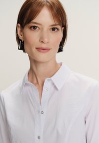 Donna con capelli corti castani che indossa orecchini triangolari neri e una camicia bianca con bottoni, di fronte con un'espressione neutra.