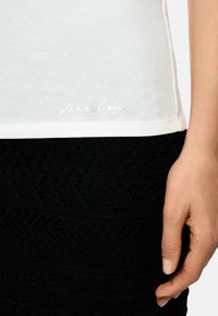 White t-shirt with a rounded hem featuring "Marc Cain" in cursive at the bottom, paired with a textured black skirt.