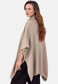 Femme aux longs cheveux bruns portant un poncho beige à col roulé ample et un pantalon foncé, regardant par-dessus son épaule.