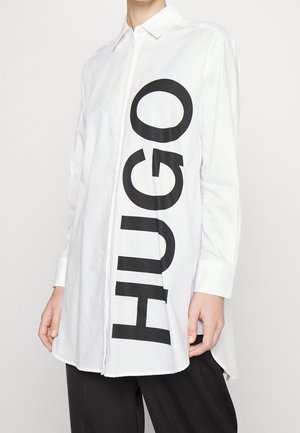 Henkilö pukeutuneena valkoiseen pitkähihainen nappipaitaan, jonka edessä on suuret pystysuorat mustat "HUGO"-kirjaimet, yhdistettynä mustiin housuihin.