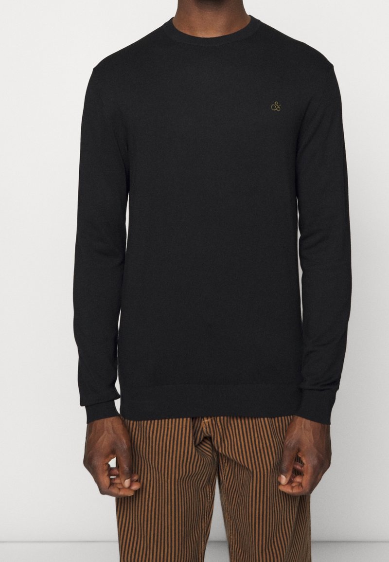 Homme portant un pull noir uni à manches longues et un pantalon marron avec des rayures verticales noires sur fond blanc.