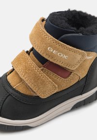 Zwarte en bruine sneaker van suede en nylon, met klittenbandsluitingen, textuuraccenten en een rubberen zool met een profielpatroon.