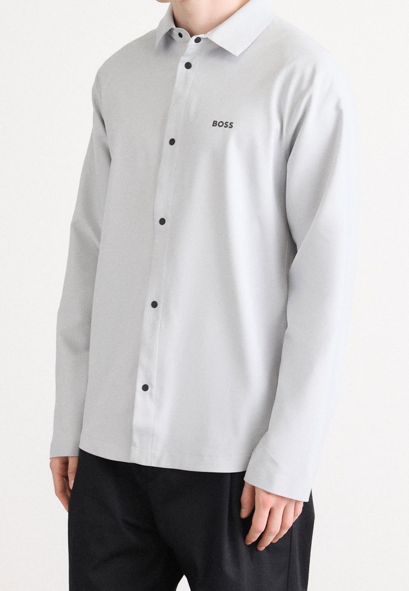 Chemise gris clair à manches longues avec des boutons noirs et le logo "BOSS" sur la poitrine, portée avec un pantalon noir sur une personne debout.