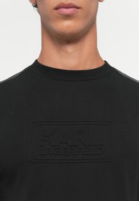 Detailný záber na osobu, ktorá má na sebe čiernu mikinu so zvýrazneným logom "Karl Lagerfeld" na hrudi a koženými detailmi na pleciach.