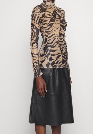 Femme portant un haut à manches longues imprimé zèbre beige et noir, associé à une jupe en cuir noire longueur genou sur un fond uni.