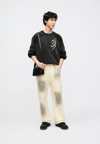 Sweatshirt preta com detalhes gráficos brancos, calças de fato de treino creme com padrões de manchas pretas e sapatos pretos sem atacadores. Mala simples de tecido.