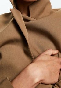 La mano di una persona che sistema il rever di un cappotto marrone chiaro con colletto alto, con attenzione alla texture del tessuto e alle pieghe.