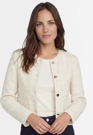 Frau mit einer cremefarbenen, strukturierten Jacke mit Knöpfen über einem weißen Top, langen braunen Haaren und neutralem Gesichtsausdruck, stehend vor einem hellen Hintergrund.