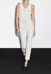 Femme marchant portant un débardeur blanc, un jean blanc taille haute, des chaussures noires et un bracelet en or à son poignet gauche.