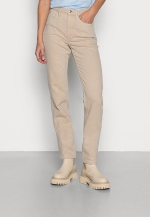 Person trägt beige Straight-Leg-Jeans, in passende klobige Knöchelstiefel gesteckt, steht vor einem schlichten hellen Hintergrund.
