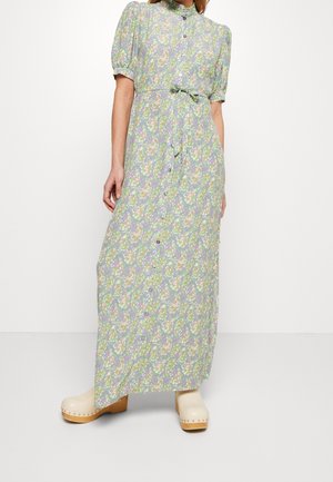 Frau trägt ein langes, geblümtes Kleid mit Knöpfen, Puffärmeln, gebundener Taille und beigefarbenen Holzclogs vor einem schlichten weißen Hintergrund.