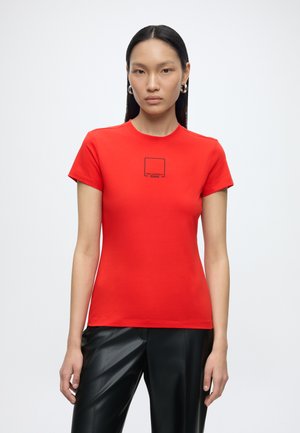 Frau mit langen schwarzen Haaren, die silberne Creolen trägt, ein rotes eng anliegendes T-Shirt mit schwarzem Text und schwarze Lederhosen, stehend vor weißem Hintergrund.