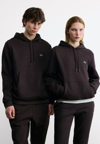 Δύο άτομα που φορούν μαύρες κουκούλες και παντελόνια που ταιριάζουν, με ένα μικρό πράσινο λογότυπο κροκοδείλου στο στήθος.