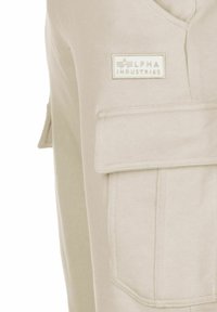 Béžová úžitková vesta s výraznou prednou kapsou a štítkom s nápisom "ALPHA INDUSTRIES". Vyrobená z mäkkej tkaniny s hladkým povrchom.
