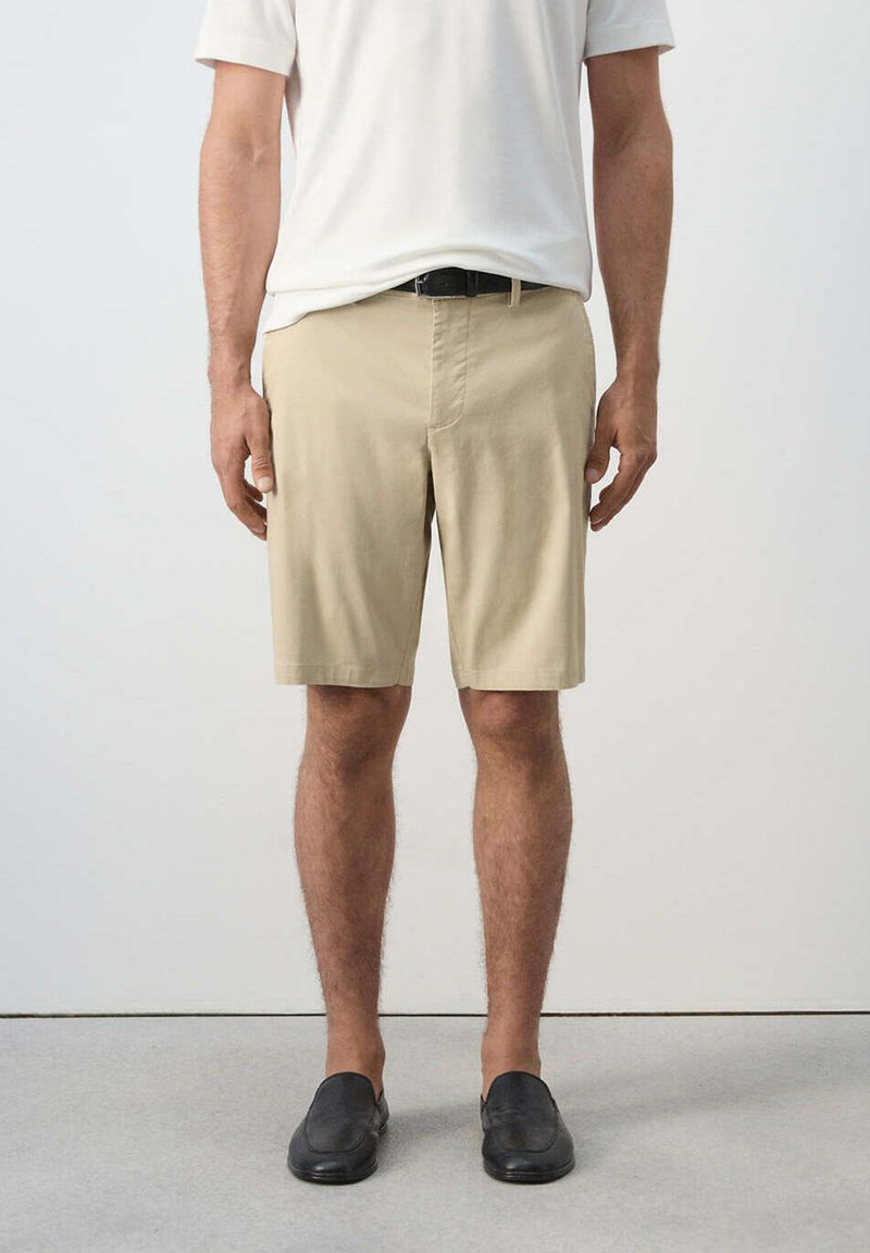 Homme portant un short beige jusqu'aux genoux, une ceinture noire, un t-shirt blanc et des chaussures noires sans lacets, debout devant un fond clair uni.